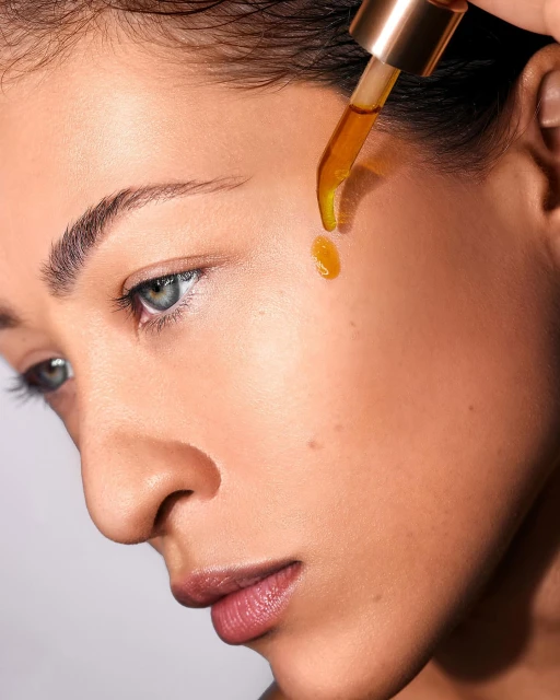 Close-up of a woman with fair skin applying golden-hued facial oil with a glass dropper, set against a soft white backdrop.