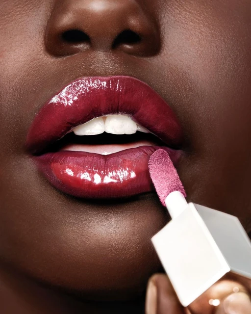 Close-up shot of an African American adult female applying glossy, burgundy lip color with a white applicator in an indoor setting.