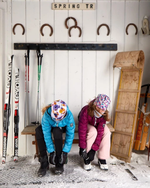 Two children in colorful winter gear sit on a bench and put on their boots inside a snowy ski lodge, with skis and sled nearby.