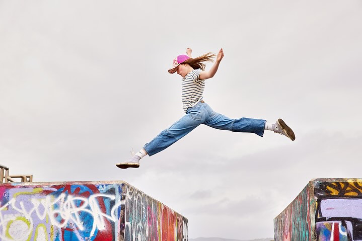 A young adult woman in mid-air, captured in a dynamic jump against a cloudy sky, wearing casual attire and a pink cap, embodying freedom and energy.