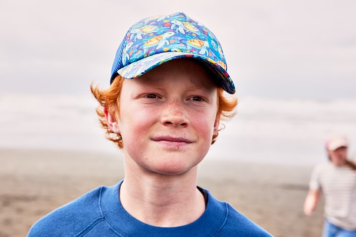 Portrait of a red-haired boy wearing a colorful baseball cap, posing against a beach backdrop with another child in the background.