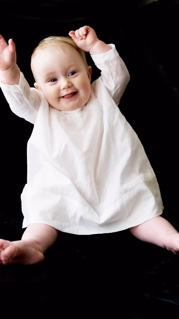 A baby in a white dress is smiling and reaching upwards against a black background. A joyful and tender moment captured.