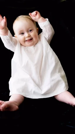 A baby in a white dress sitting against a black background with hands raised.