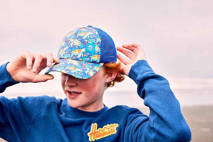 Young person wearing a Turtle Fun baseball cap with sea turtle pattern, adjusting the brim in an outdoor environment on a cloudy day.