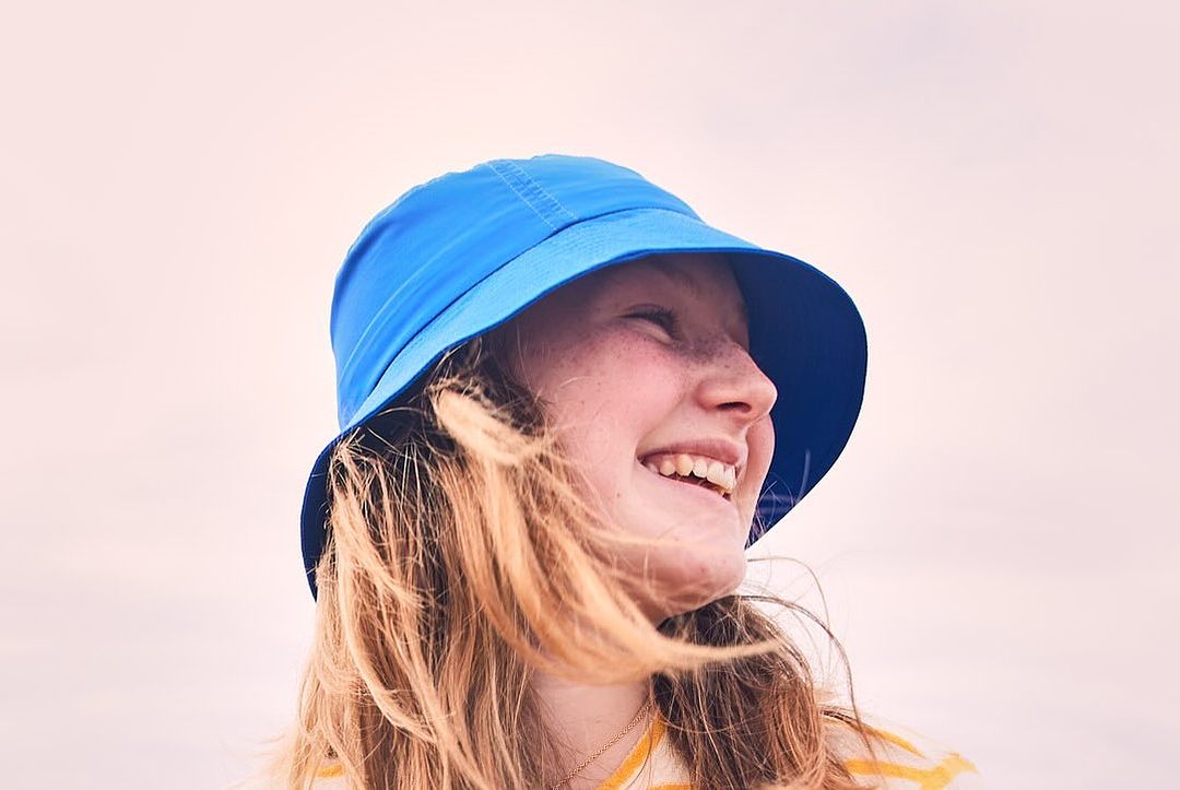 Profile of a smiling young woman with freckles wearing a blue bucket hat outdoors, highlighted by a light, airy backdrop.