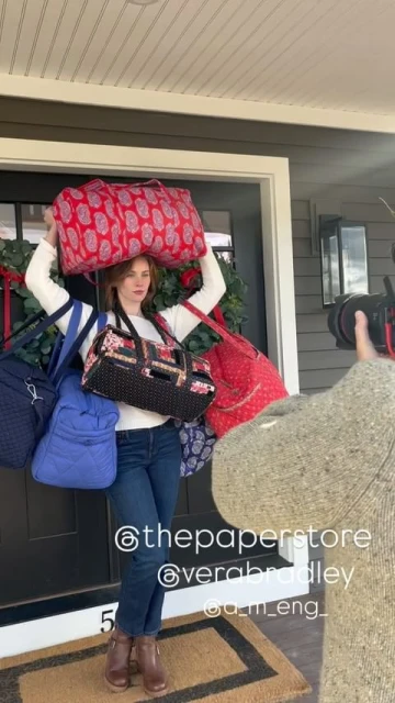 Woman posing with variety of Vera Bradley bags near a front door, with a photographer partially visible on the right.