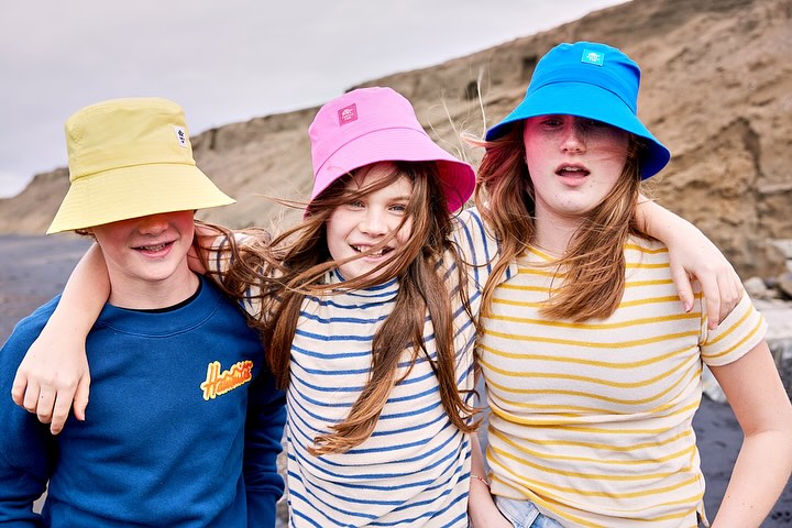 Three children smile outdoors on a beach with a rocky hill backdrop. They wear colorful bucket hats and striped shirts in a medium shot.