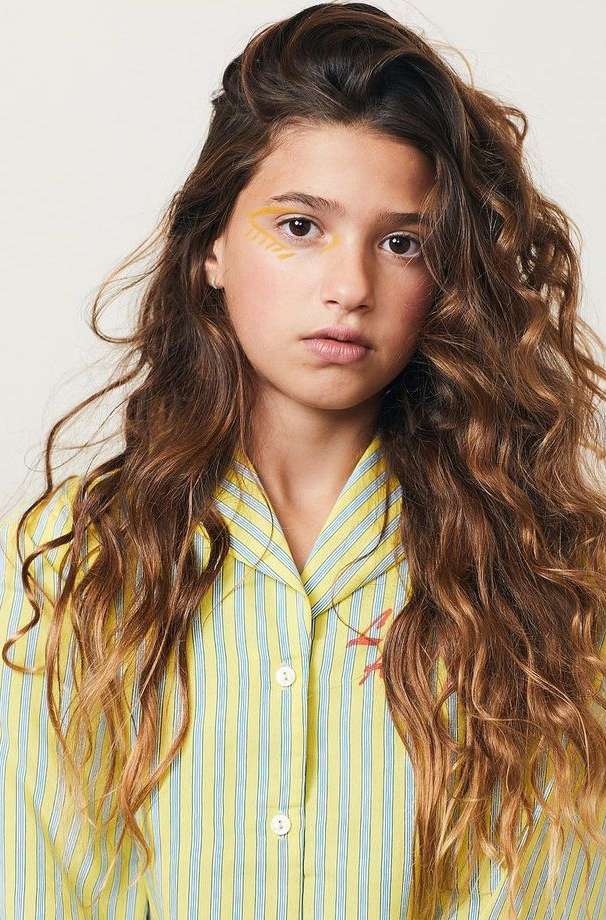 Portrait of a young female with light brown wavy hair, wearing a yellow and blue striped pajama top with yellow face paint.