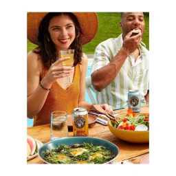 A smiling woman in an orange top and wide-brimmed hat holds a wine glass outdoors, with a man eating beside her at a wooden table.