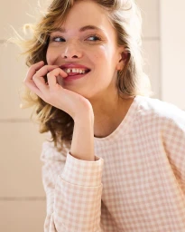 Close-up portrait of a smiling woman in a pink and white gingham sweatshirt biting her fingernail