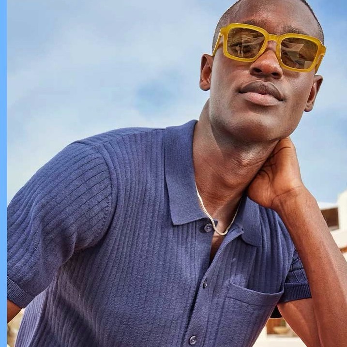 A dark-skinned man wears a blue polo shirt and yellow sunglasses against a blue sky, looking at the camera with a neutral expression.