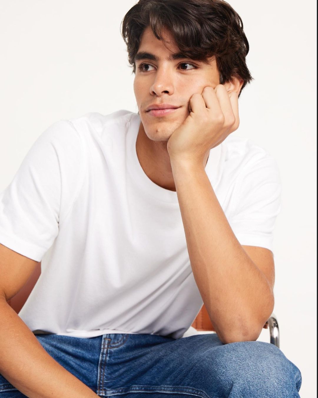 Portrait of a young adult male in a white t-shirt and jeans, sitting against a white backdrop, looking to his left.