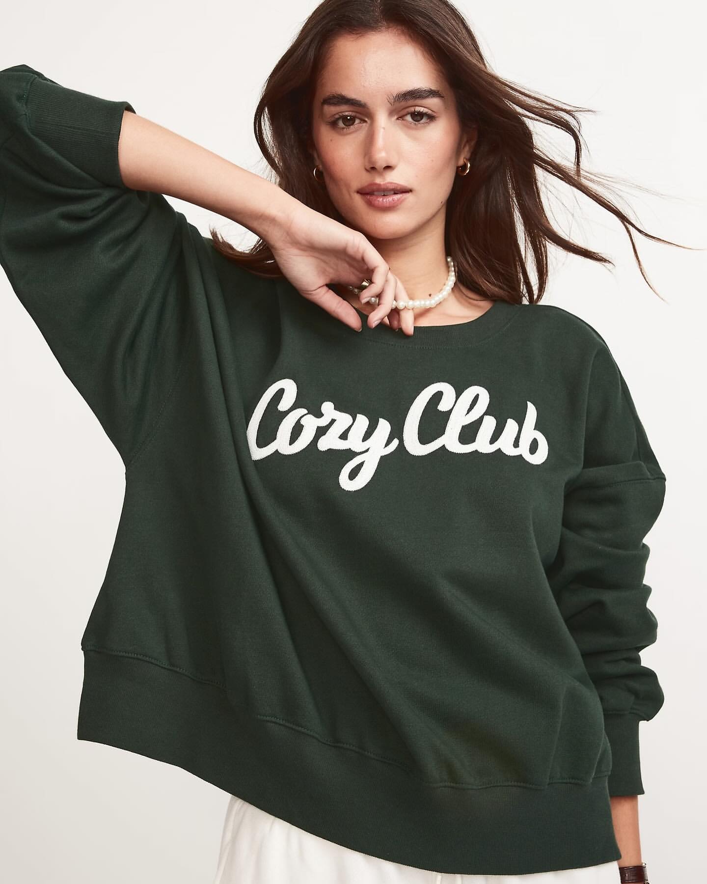 A woman poses in a dark green 'Cozy Club' sweatshirt, pearl necklace, and light pants against an off-white backdrop in a studio shot.