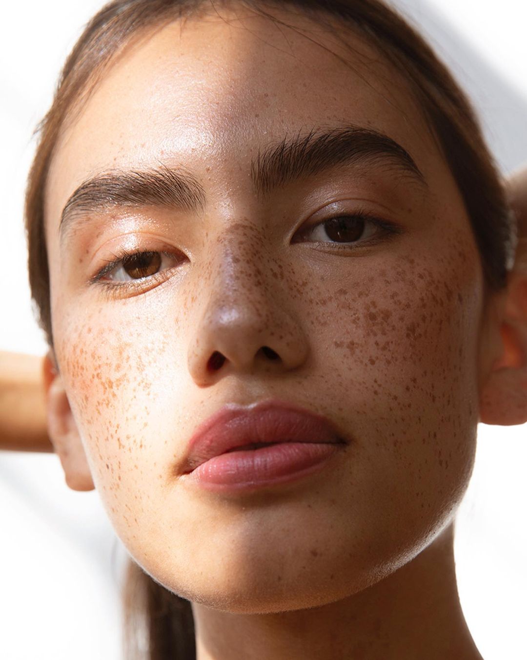 Close-up studio shot of a woman with freckles, tan skin, and glossy lips. The subject has her hands behind her head and wears no visible clothing.