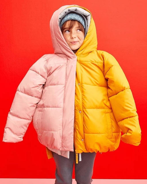 Child in a vibrant two-tone pink and yellow puffer jacket, smiling indoors against a red background, wearing a blue knit hat.