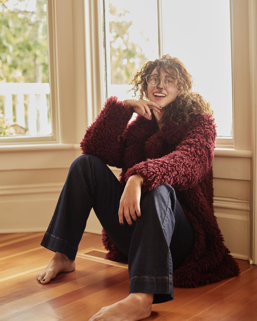 Woman with curly hair, wearing glasses and a fuzzy jacket, sits barefoot on a wood floor near a window with greenery outside.