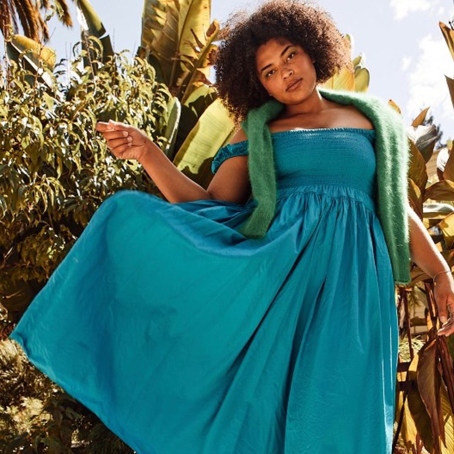 Young woman with afro wearing turquoise dress and green shawl posing in front of tropical foliage in outdoor setting during daytime.