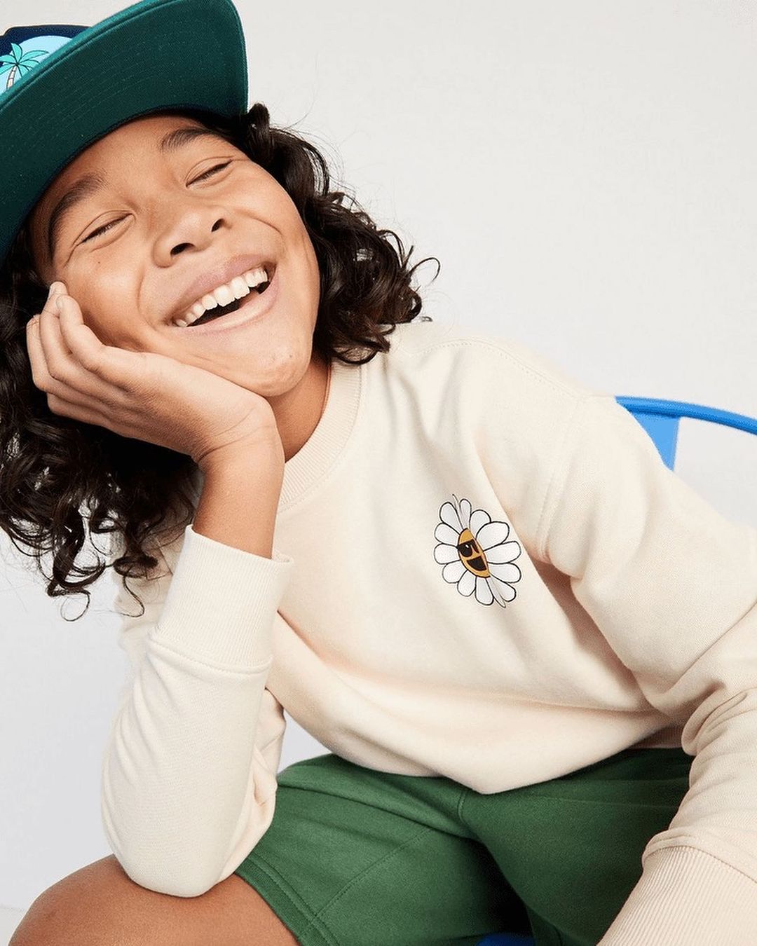 Smiling young boy wearing a baseball cap and sweatshirt with a daisy graphic sits on a chair indoors in a close-up shot.