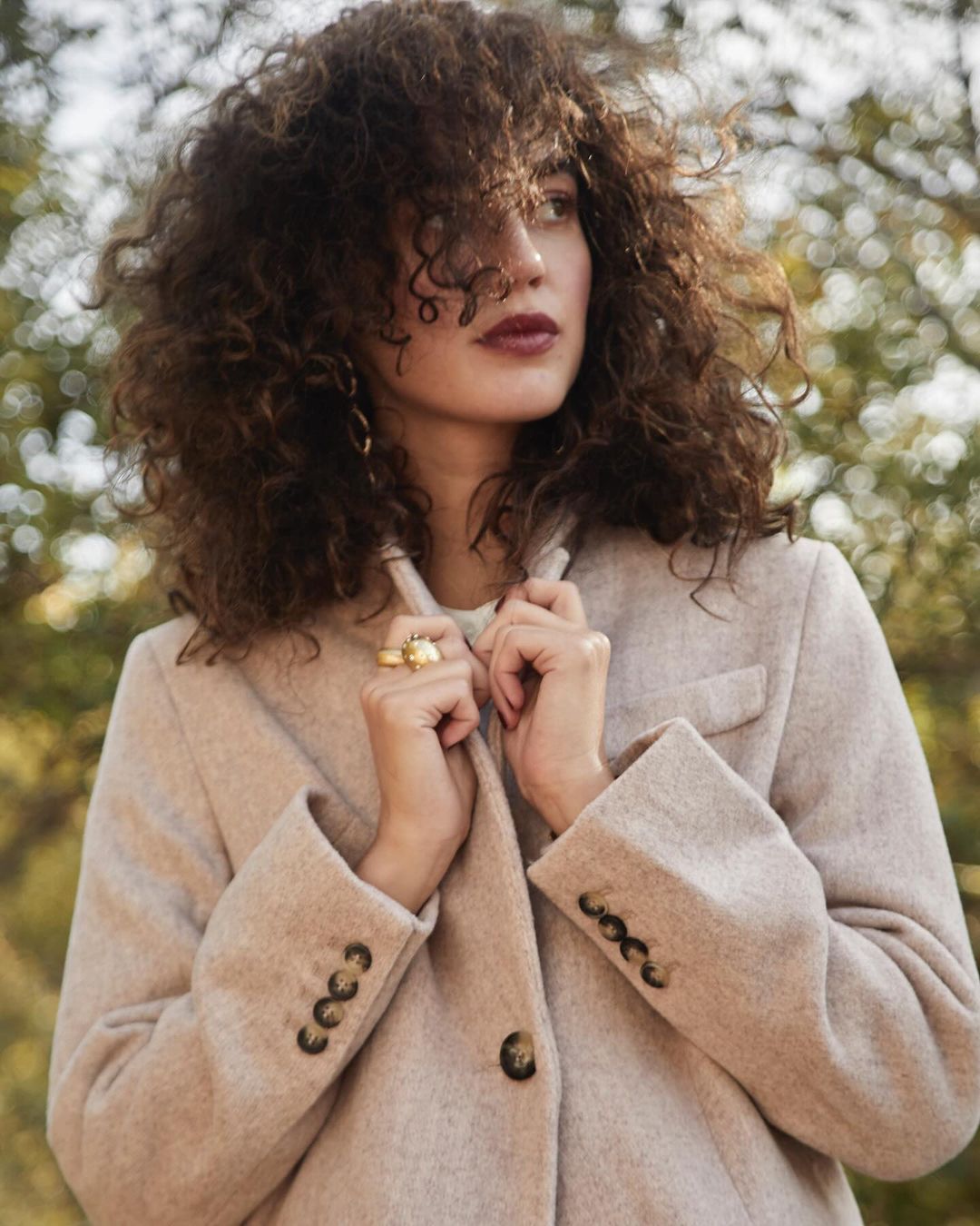 A woman with curly hair wearing a light-colored coat stands outdoors. The photo features a shallow depth of field with soft, blurred background.