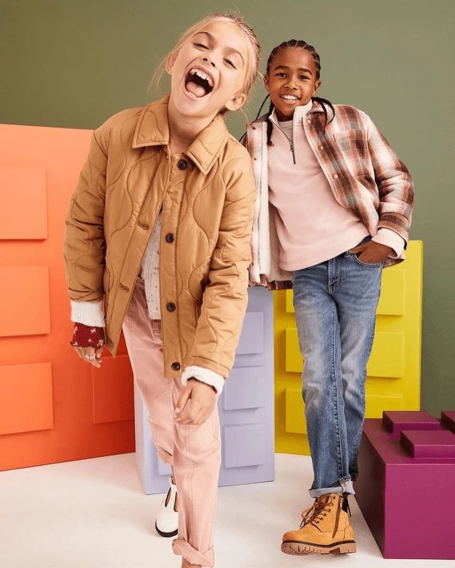 Two smiling kids pose in a bright studio. One wears a tan jacket and pink pants; the other wears a flannel shirt and blue jeans.