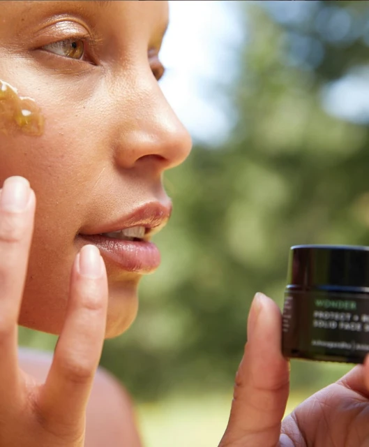 Adult female applies facial mask with trees in the background. Skincare product in foreground.
