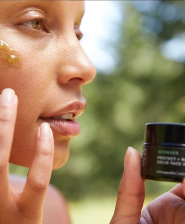 Adult female applies facial mask with trees in the background. Skincare product in foreground.
