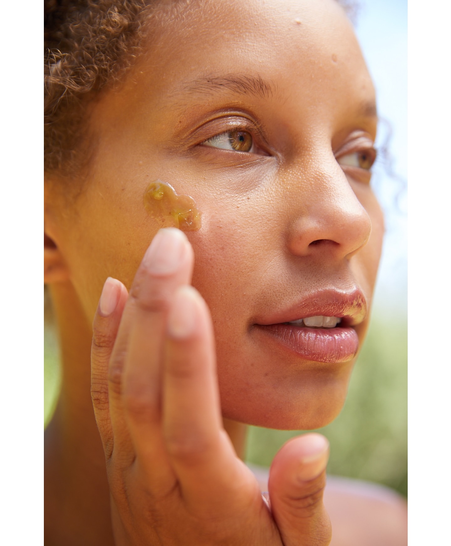 Close-up portrait of a woman applying golden serum to her face outdoors, with a shallow depth of field and natural lighting.