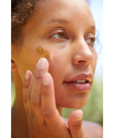 Close-up portrait of a woman applying golden serum to her face outdoors, with a shallow depth of field and natural lighting.