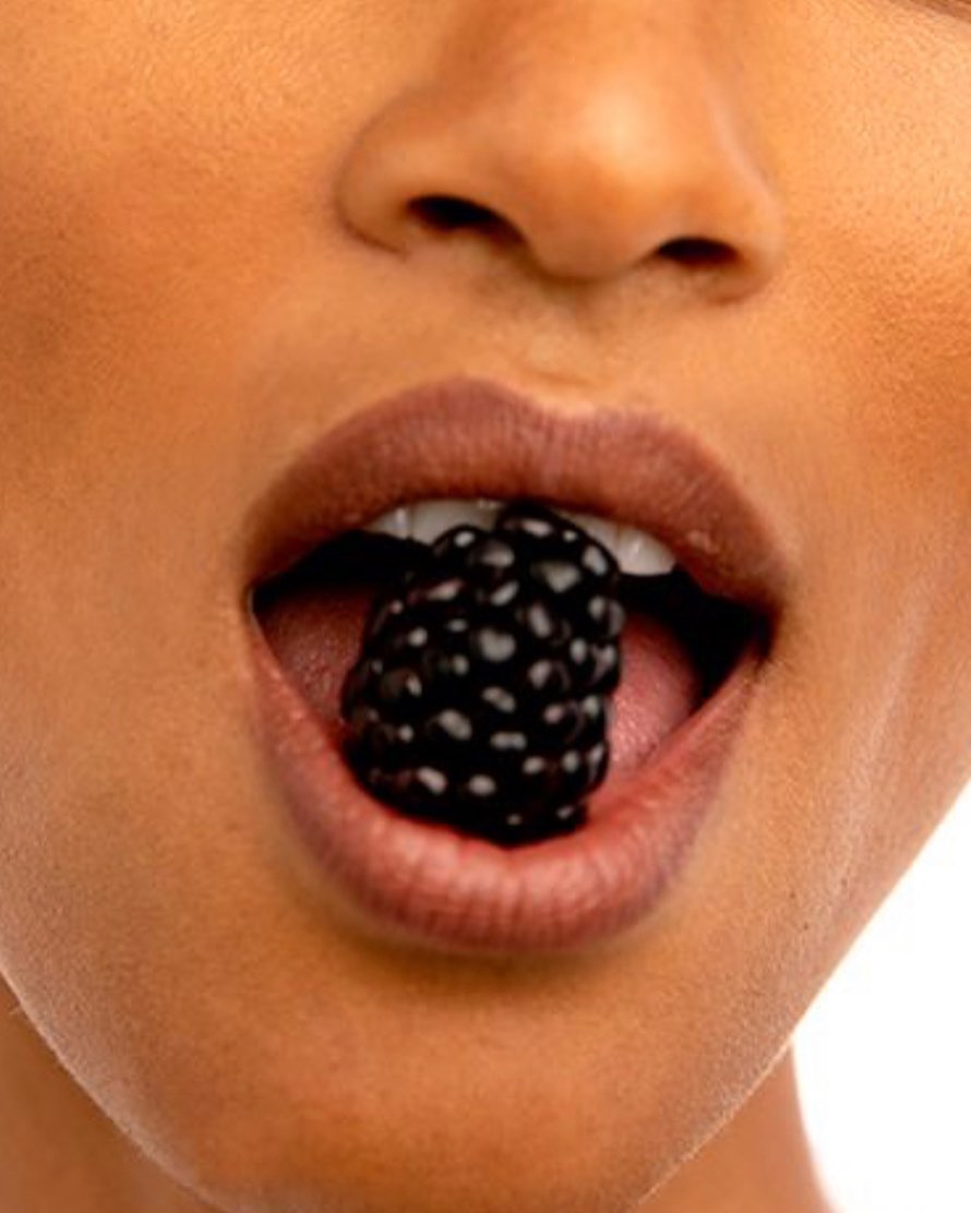 Close-up of an adult woman's face with tan skin eating a blackberry, set against a white background in a studio food shot.