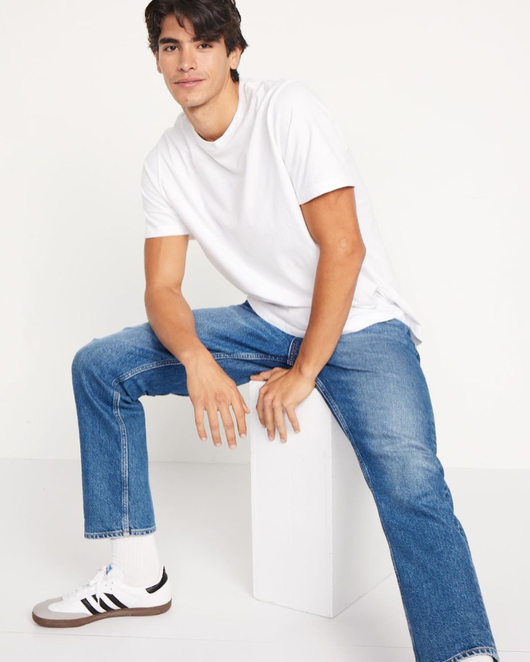 A young man sits in a casual pose wearing a white t-shirt and blue jeans with sneakers, against a plain neutral background.