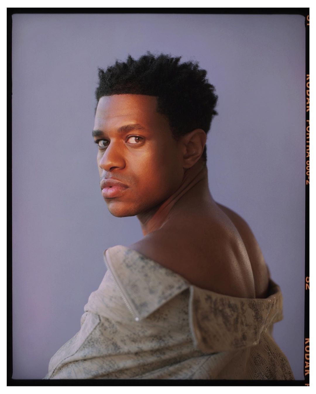 Portrait of a dark-skinned adult male with an off-the-shoulder jacket in a studio setting with soft lighting and neutral background.