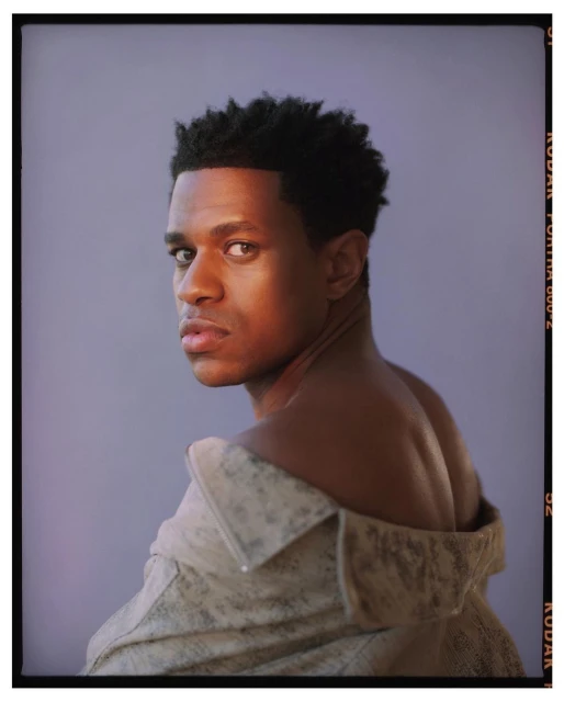 Portrait of a dark-skinned adult male with an off-the-shoulder jacket in a studio setting with soft lighting and neutral background.