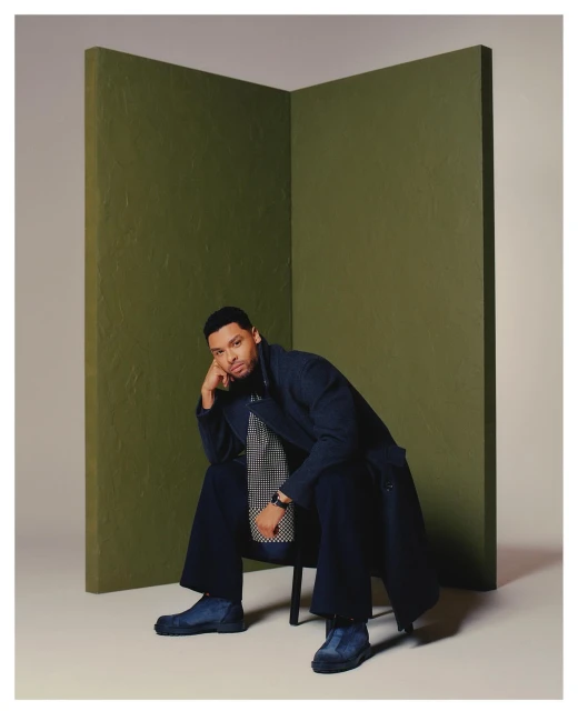 Portrait of a seated adult male wearing a navy overcoat and trousers, with an olive green backdrop in a studio environment.