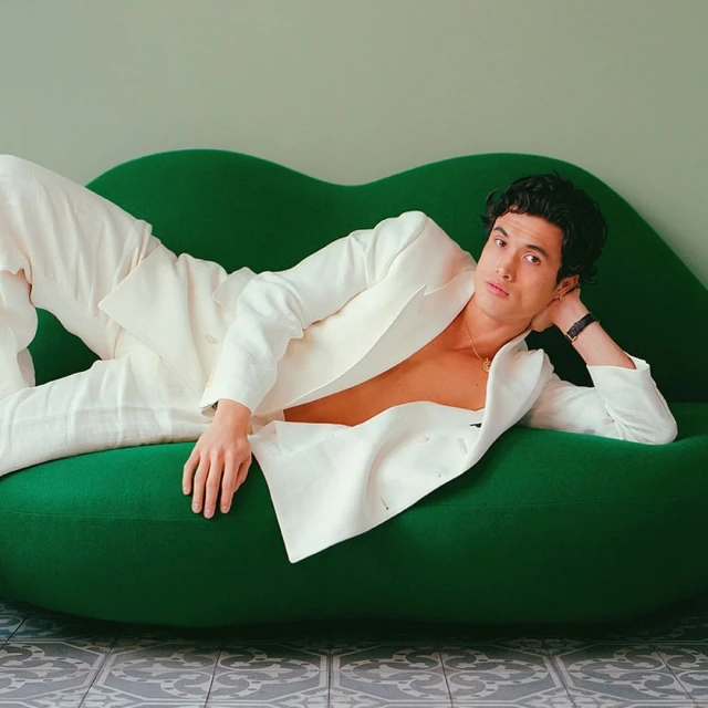 Adult male in a white suit poses while reclining on a green velvet couch in an indoor setting with a tiled floor and neutral wall.