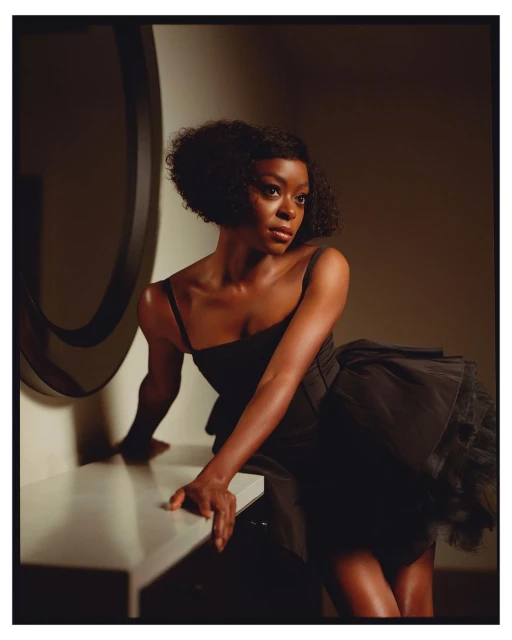 Elegant studio portrait of a young Black woman with a short Afro hairstyle, wearing a black dress, leaning on a white surface with a round mirror.