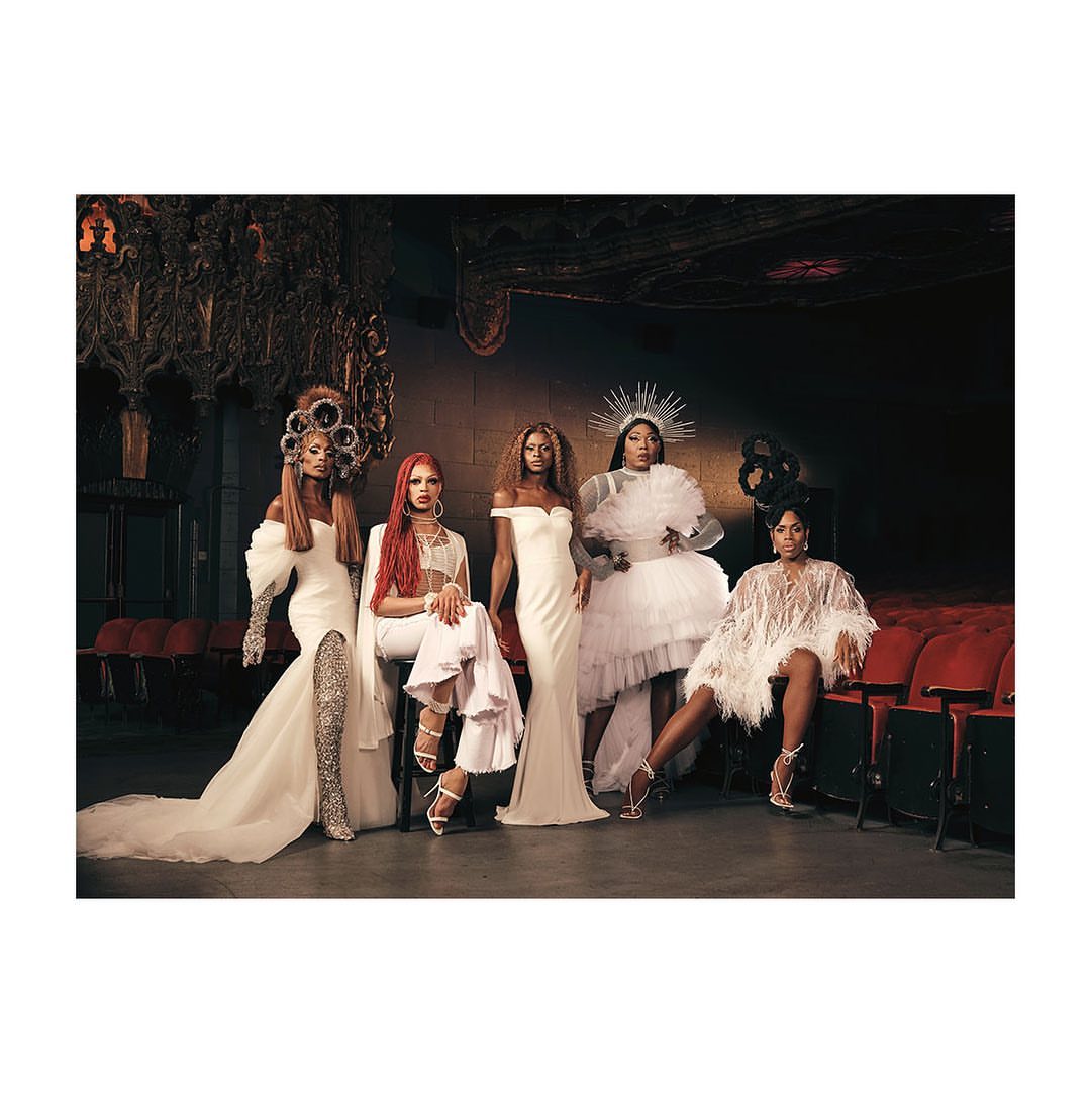 Five drag queens pose in an ornate theatre in all-white gowns, showcasing a blend of glamour, artistry, and theatrical atmosphere.