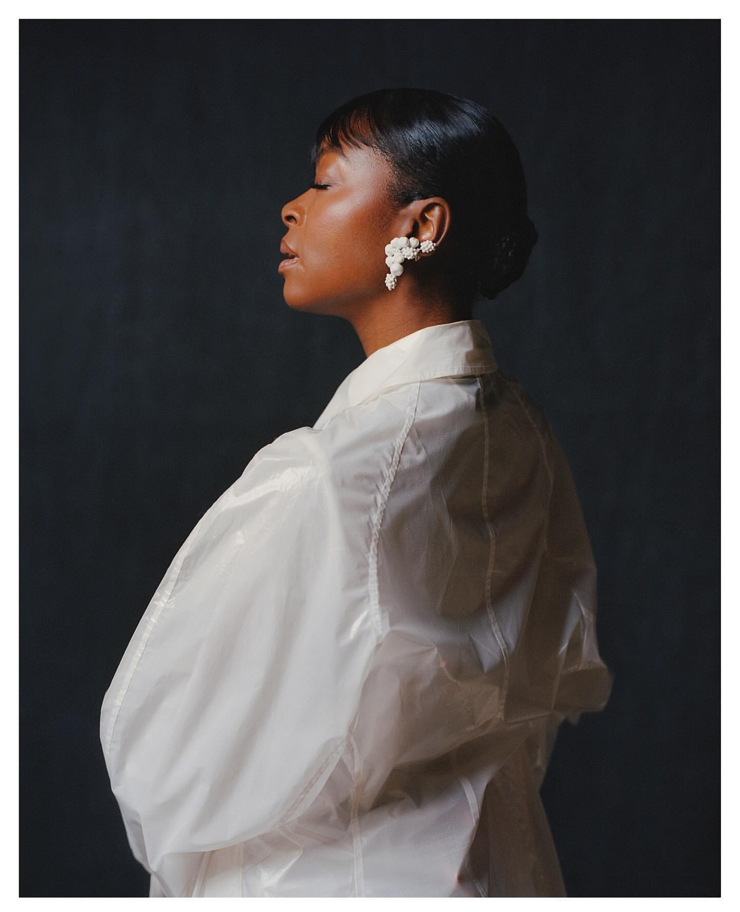Elegant portrait of a woman in a translucent white jacket and floral earrings, captured against a dark gray backdrop with soft lighting.