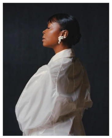 Elegant portrait of a woman in a translucent white jacket and floral earrings, captured against a dark gray backdrop with soft lighting.