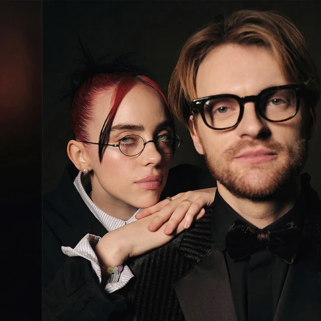 Portrait of siblings Billie Eilish and Finneas O'Connell posing together indoors wearing formal attire in front of a dark background.
