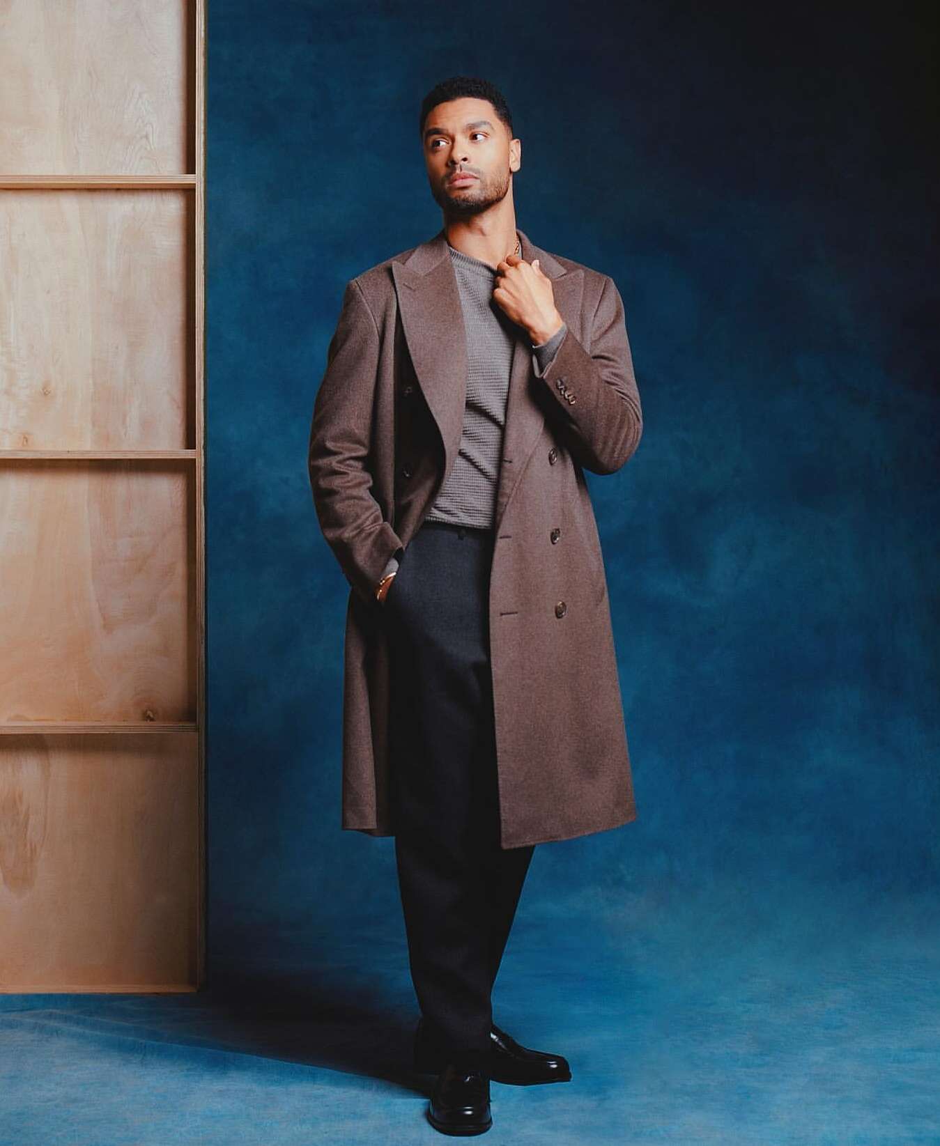 Portrait of Regé-Jean Page wearing a brown overcoat against a dark blue textured backdrop with a wooden frame structure.