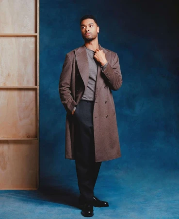 Portrait of Regé-Jean Page wearing a brown overcoat against a dark blue textured backdrop with a wooden frame structure.