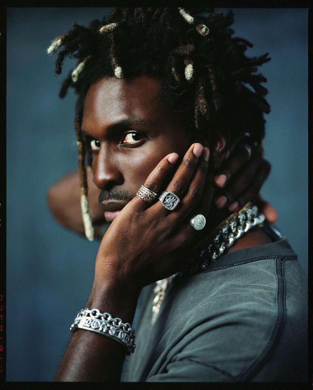 Close-up portrait of a black adult male with dreadlocks, wearing a gray shirt and multiple rings and bracelets, against a grey background.