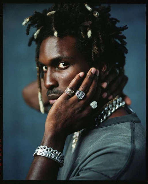 Close-up portrait of a black adult male with dreadlocks, wearing a gray shirt and multiple rings and bracelets, against a grey background.