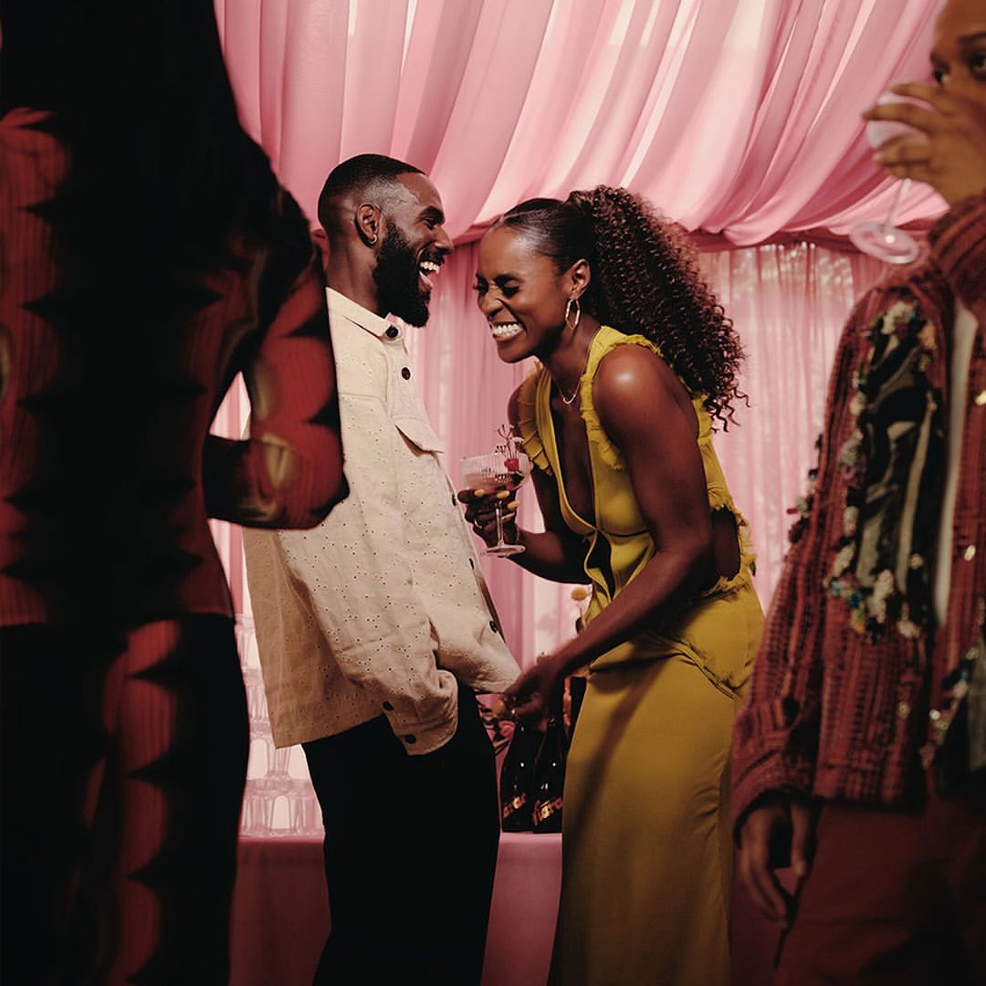 A happy couple laughs together indoors at an event with other people. The man wears a beige jacket; the woman wears a yellow dress.