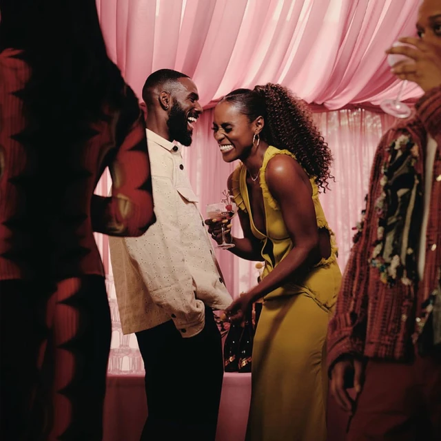 A happy couple laughs together indoors at an event with other people. The man wears a beige jacket; the woman wears a yellow dress.