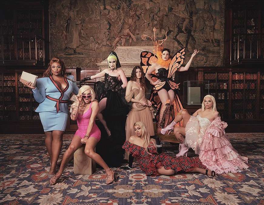 A diverse group of drag queens in gowns and dresses strike a fashionable pose in a library with books and tapestries in the background.