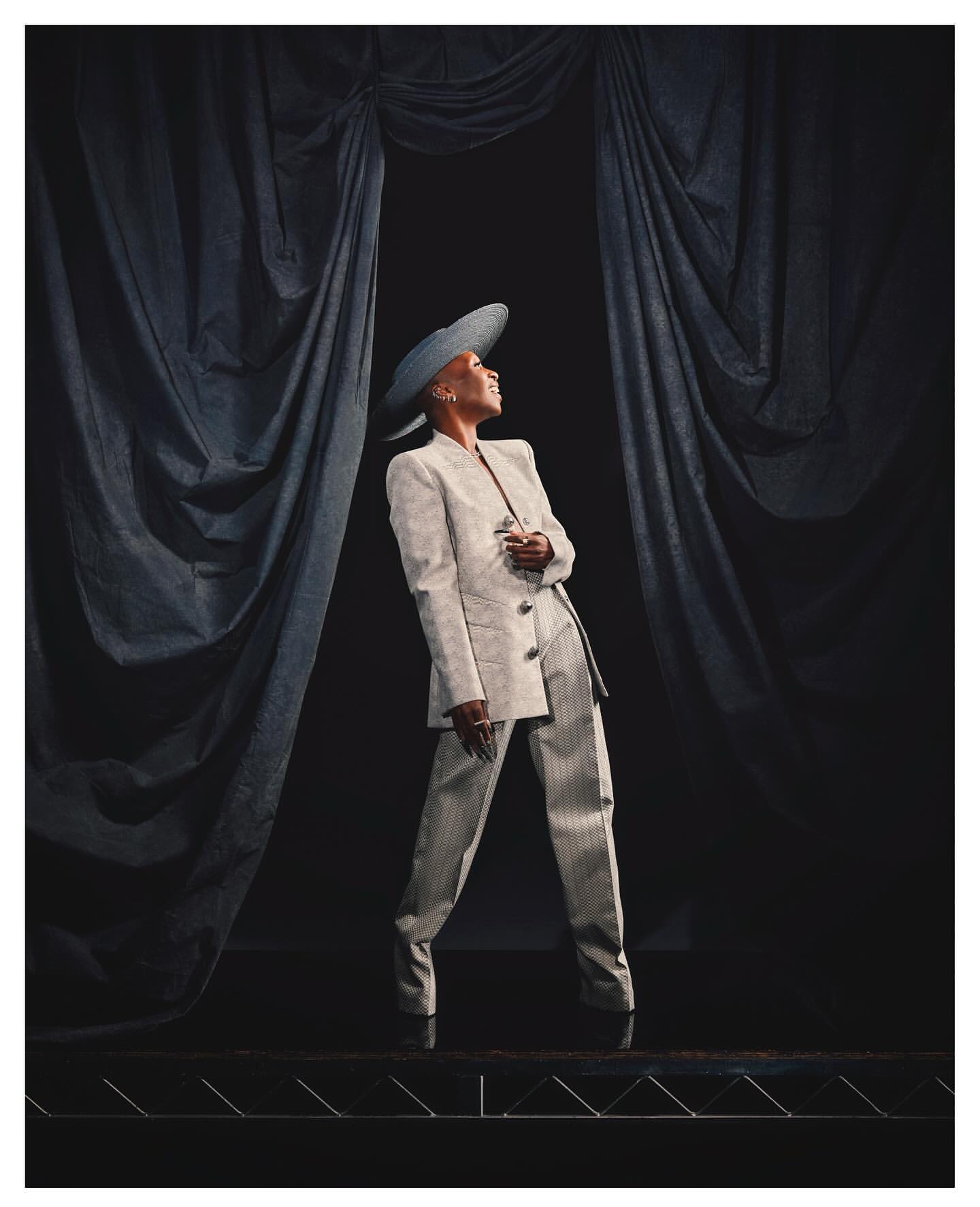 Elegant black woman in a cream suit and hat strikes a pose in front of dark drapes, showcasing fashion and poise under soft light.