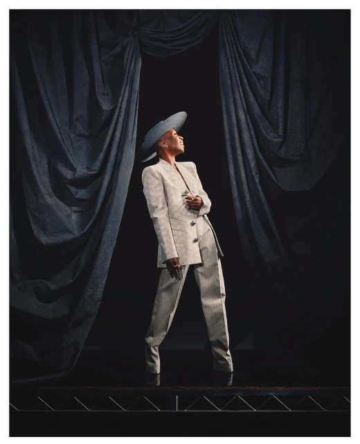 Elegant black woman in a cream suit and hat strikes a pose in front of dark drapes, showcasing fashion and poise under soft light.