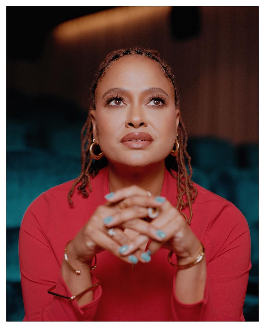 Portrait of a woman with brown locs wearing a red suit jacket, gold hoop earrings, and bracelets, looking up with her hands clasped.