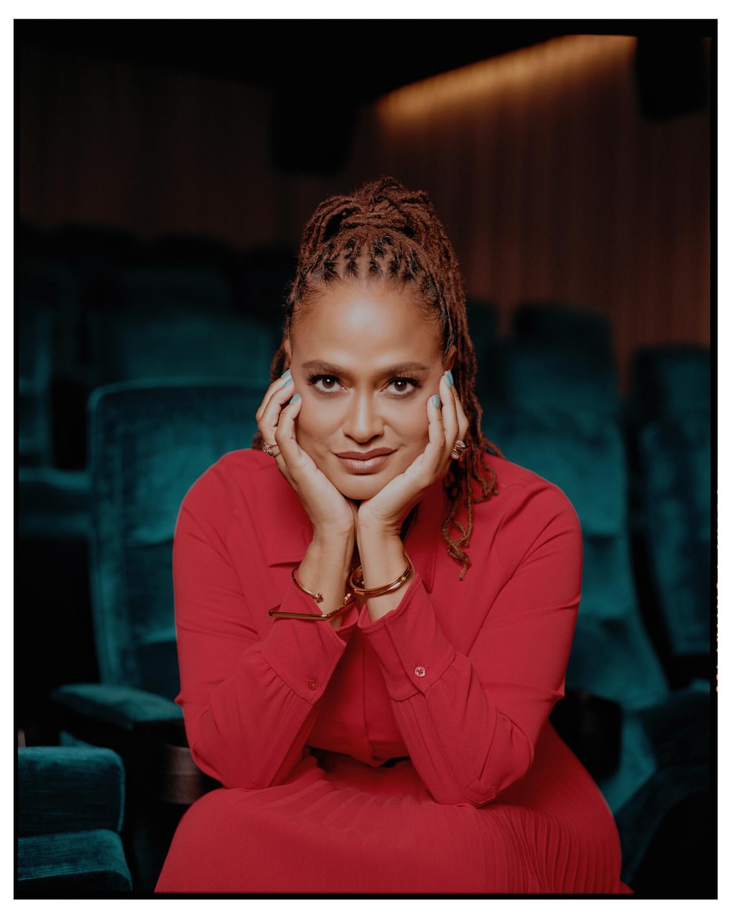 A woman with braided hair, wearing a red dress, is seated in a theatre with her hands near her face, smiling in a medium shot.